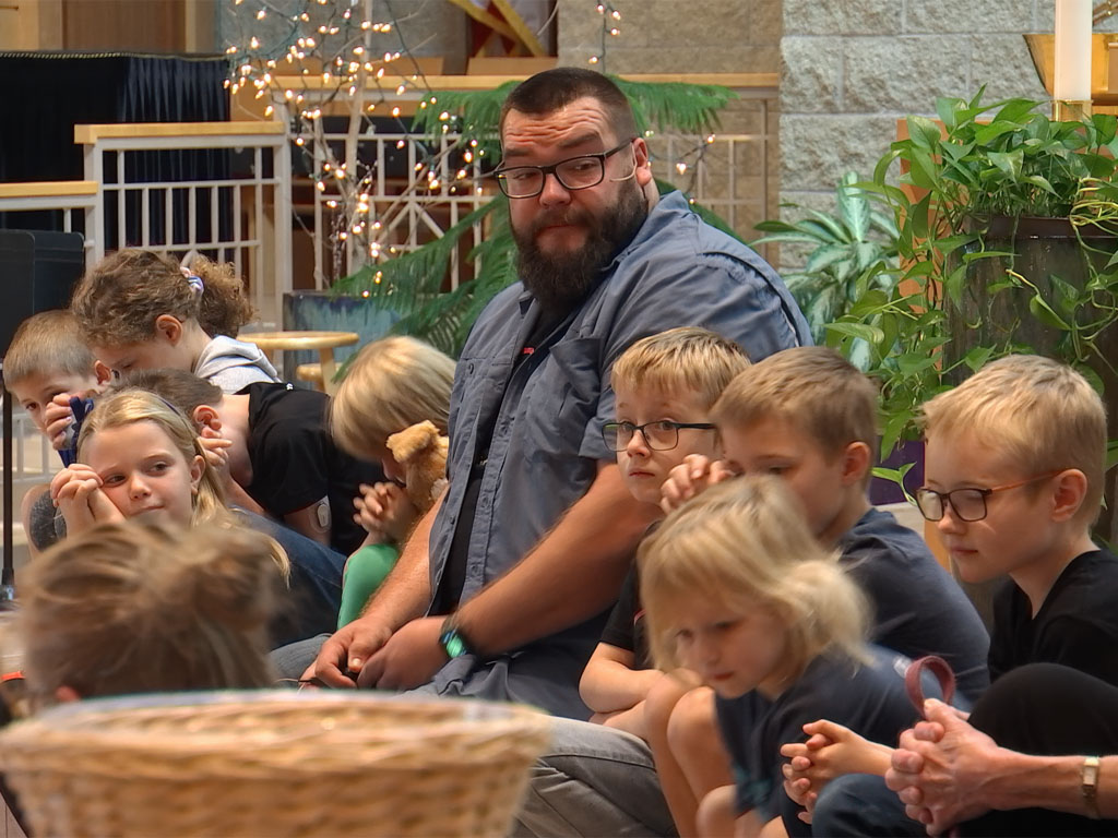 Man with a beard wearing a wireless microphone sits among a group of children at the front of a church.