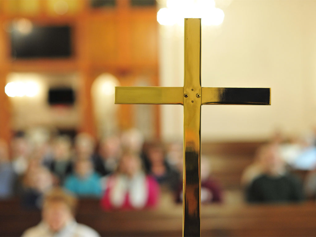 A thin, brass cross sits on an altar in front of an out-of-focus group of congregants in church pews.  