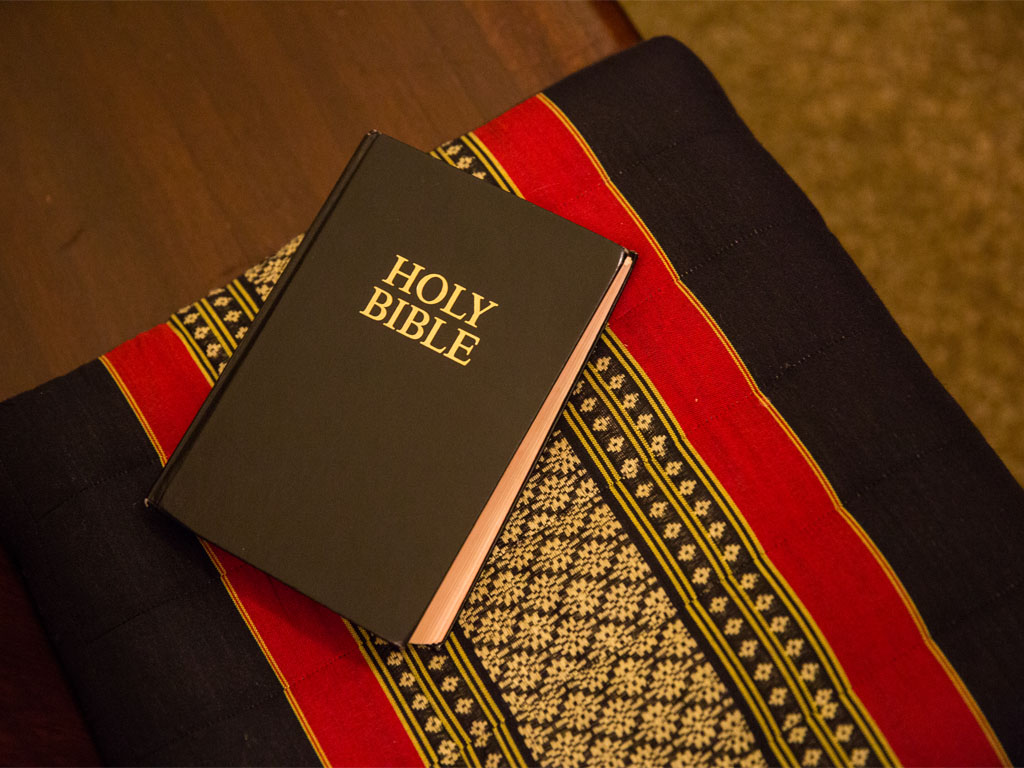 view from above of a dark brown book with HOLY BIBLE embossed in yellow on the front sitting atop a table covered with a stitched table runner with black, red, and yellow strips of fabric.