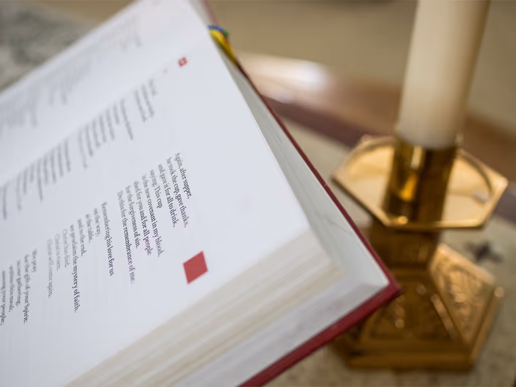 red worship book open to a page of the liturgy focused on communion sitting in front of an out-of-focus brass base holding a white candle.
