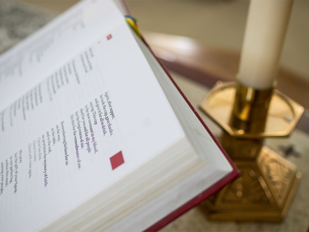 red worship book open to a page of the liturgy focused on communion sitting in front of an out-of-focus brass base holding a white candle.