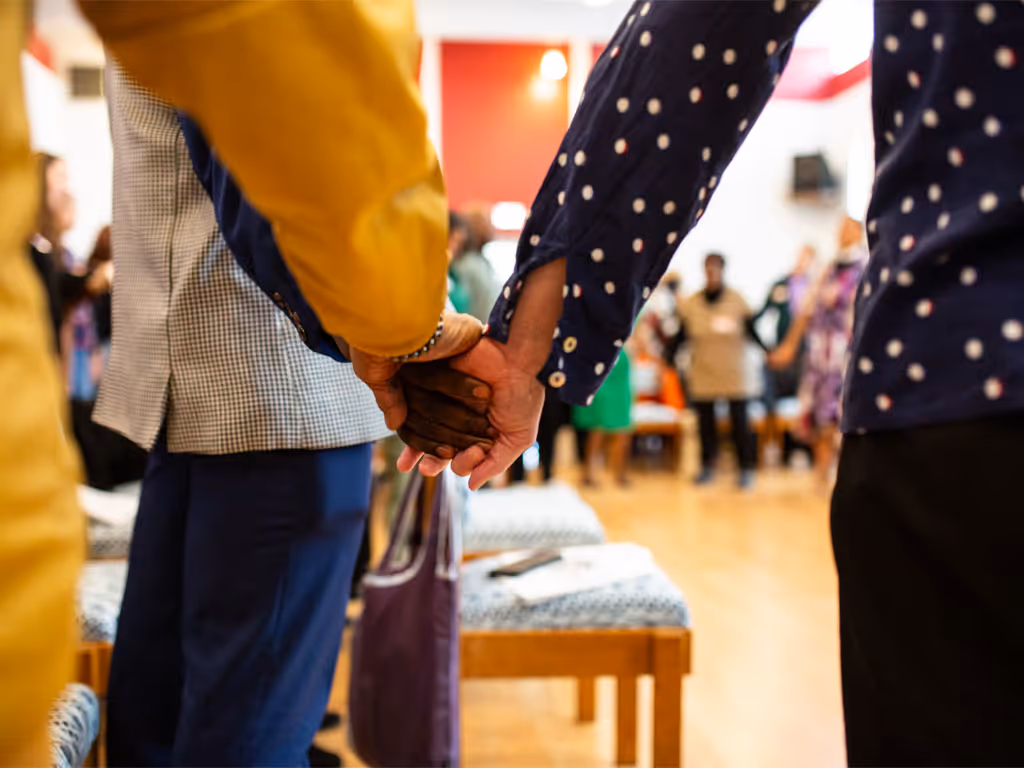 close-up of three hands joined together in prayer with a blurred view of a circle of people holding hands visible between their arms and bodies.