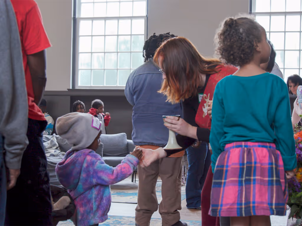Female pastor in red vestment leans over and hands a young child in a purple coat and gray hat a piece of communion bread.