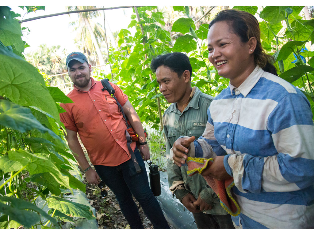 bearded man in red shirt holding a water bottle stands next to a man in a green jacket and a woman wearing a blue and white striped shirt between rows of tall plants in Cambodia.  
