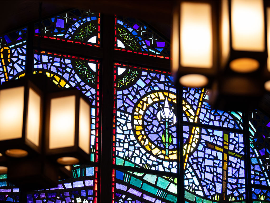 stained glass cross in black with red outline behind two blurred light fixtures with rectangular wooden frames and yellow glass. 