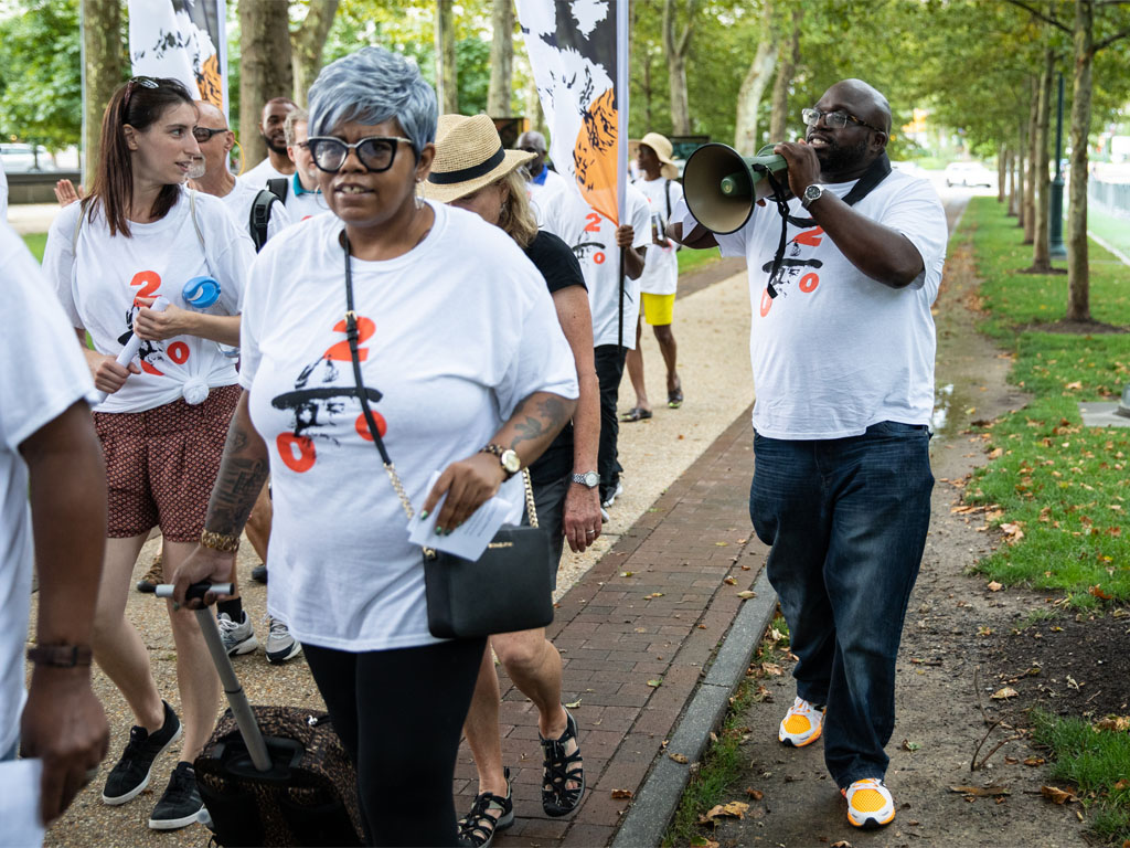 A group of people wearing white shirts, with a man’s face and the numbers 2, 0 and 0 in red, walk along a tree-lined sidewalk as a man in a matching shirt speaks through a bullhorn.