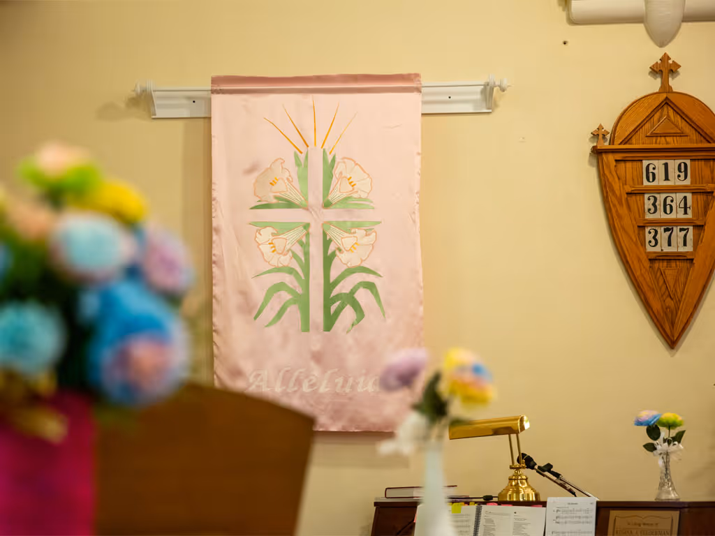 A pink banner featuring a cross outlined by palms hangs on a wall next to a wooden sign with hymn numbers. An out of-focus bouquet of flowers in blue and yellow sits in the left foreground next to the banner. 