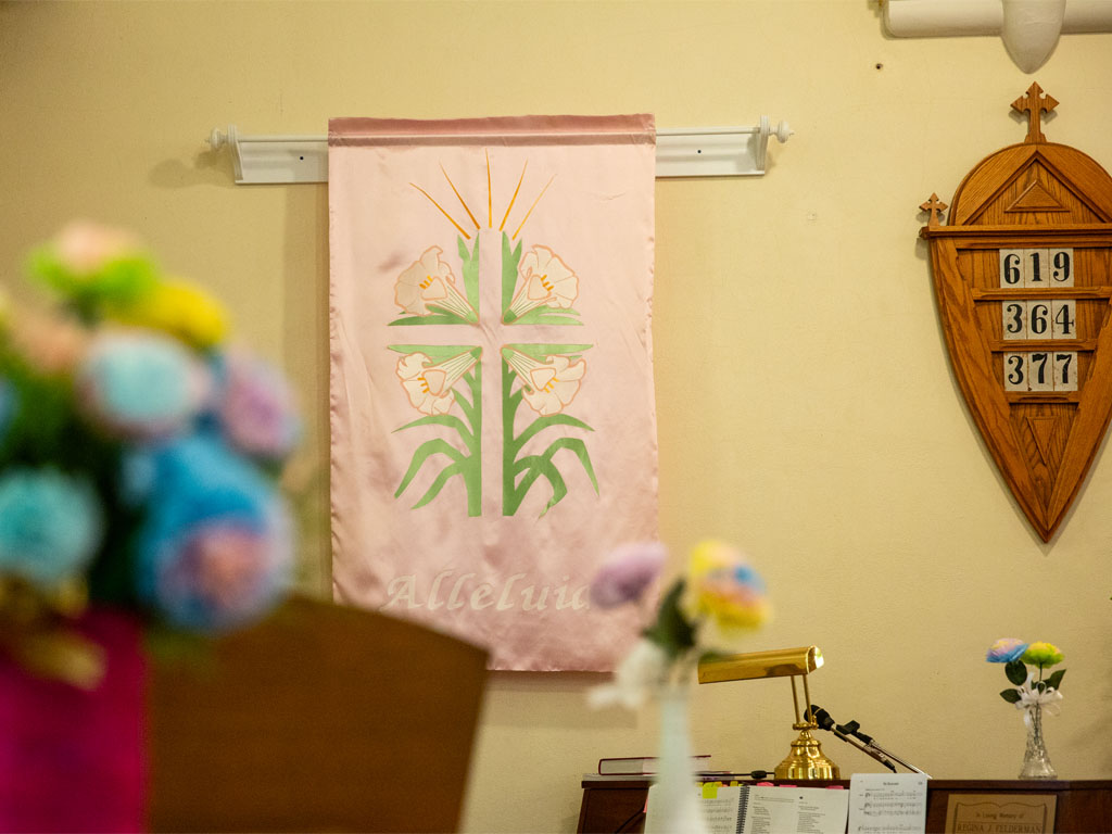 Pink banner with an image of green palms arranged into a cross hangs on a pale yellow wall next to a wooden board with three hymn numbers on it. 