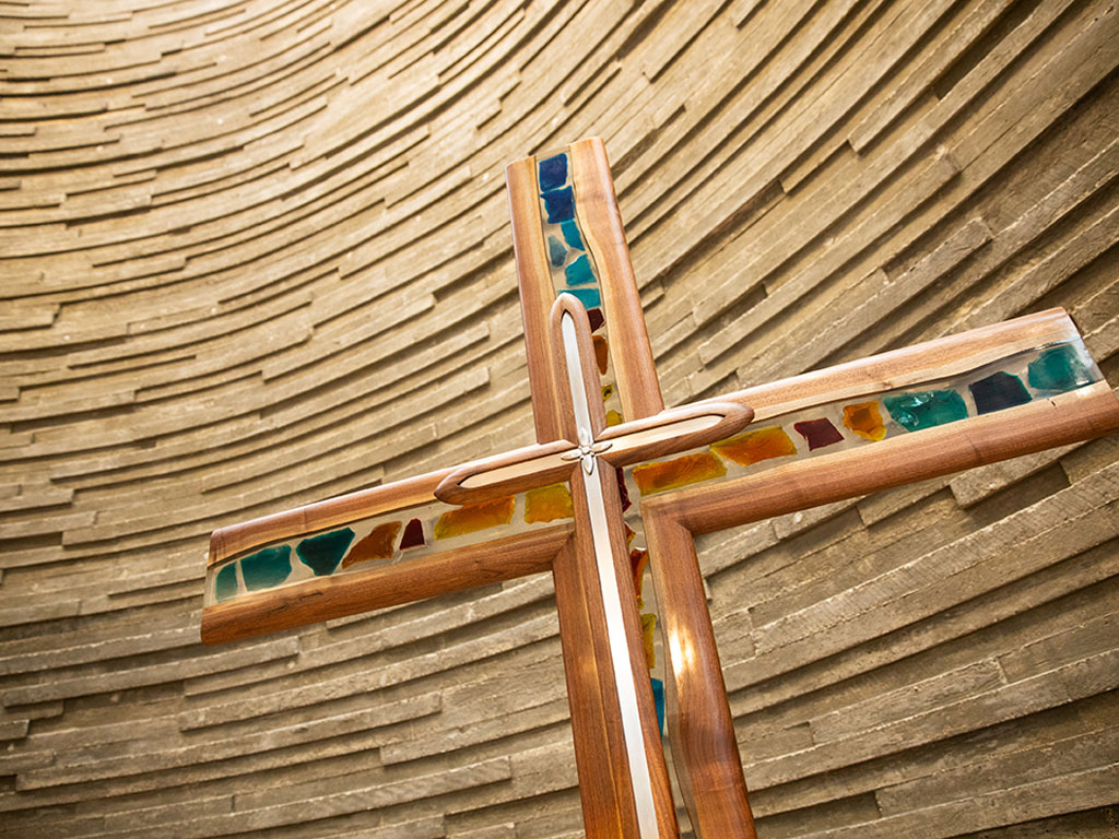 wooden cross with inlaid mosaic tiles in front of a circular wall slats of wood that are uneven