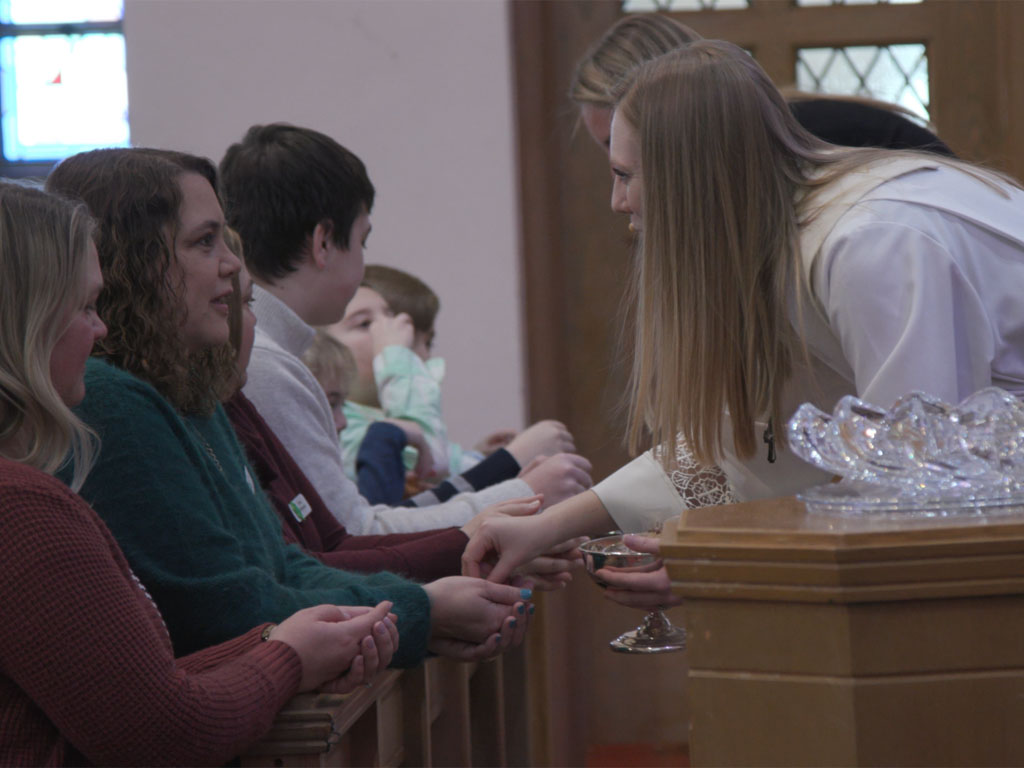 Person distributing communion to people seated at an altar rail during a church service