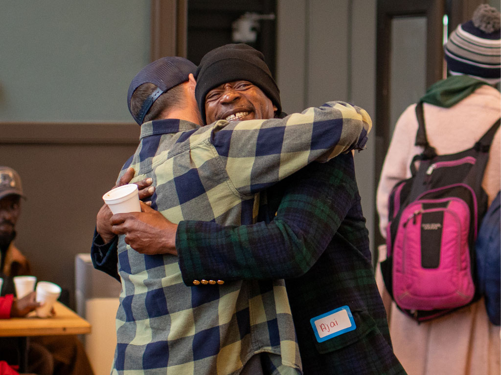 Two men wearing flannel share a hug with one man looking toward the camera, eyes closed holding a coffee cup behind the second man who is wearing a blue hat and looking away from the camera. 