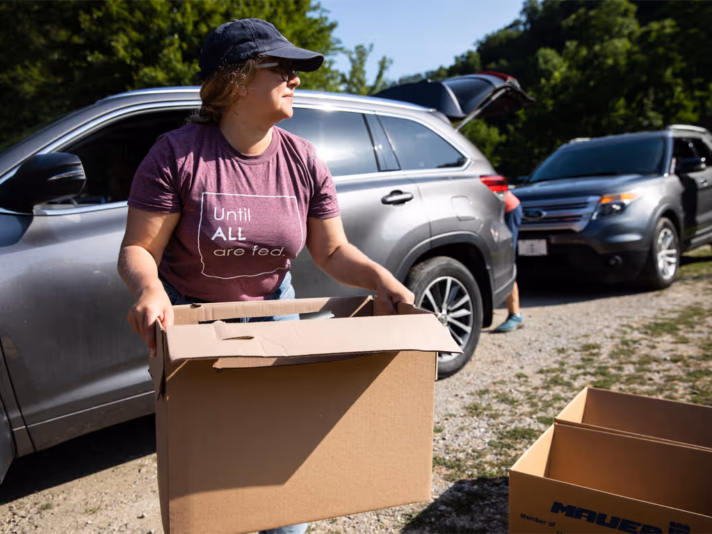 Female wearing a baseball hat and purple shirt that reads "until all are fed" carries a cardboard box from a silver SUV parked behind her.  