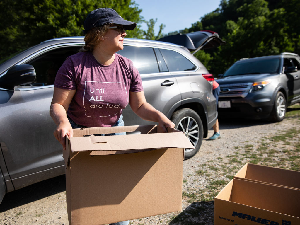 Female wearing a baseball hat and purple shirt that reads "until all are fed" carries a cardboard box from a silver SUV parked behind her.  