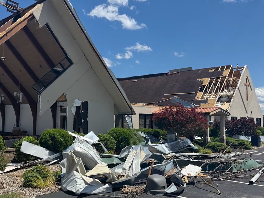 Debris sits in a pile in a parking lot in front of a church building that shows noticeable damage including a portion of the roof missing.  
