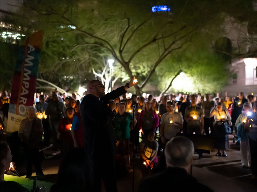 A bald man wearing a suit coat holds a tea light with his left hand as he speaks to a crowd in front of a banner that reads “AMMPARO.”  