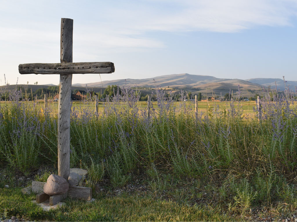 A wooden cross made from thick pieces of wood with holes poked throughout stands in front of a field of lavender with mountains in the background.