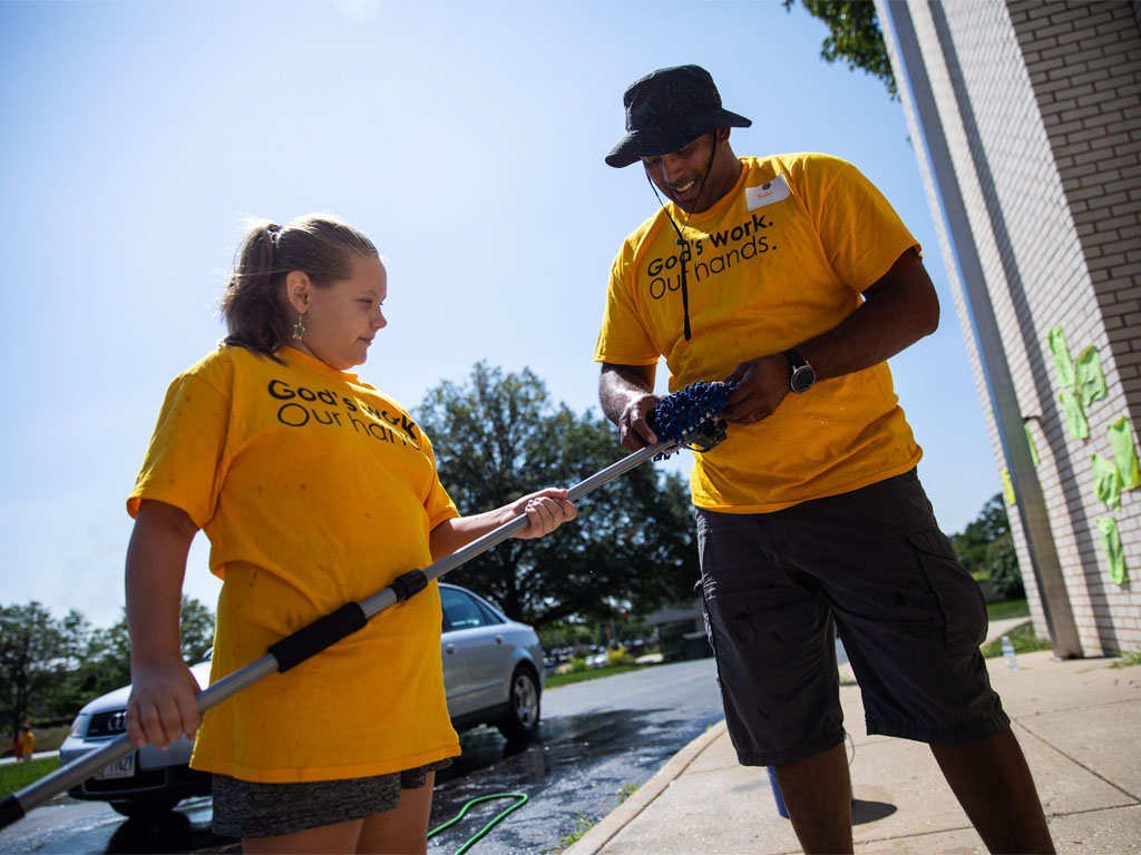 A teenage girl holds a metal pole in front of a car that is entering a car wash while a man attaches a sponge to the end of the pole. Both are wearing yellow t-shirts with the words “God’s Work. Our Hands” on them.