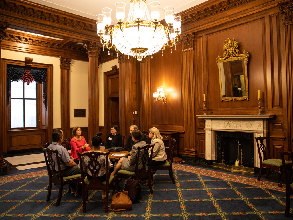 A group in formal attire sits around a circular table in a large room with high, orange wooden walls and a large chandelier. 