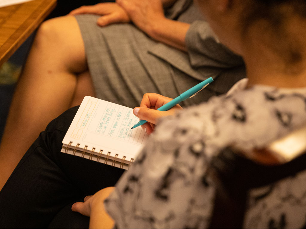 A person writes in a spiral-bound notebook with a blue pen as they sit at a round wooden table. 
