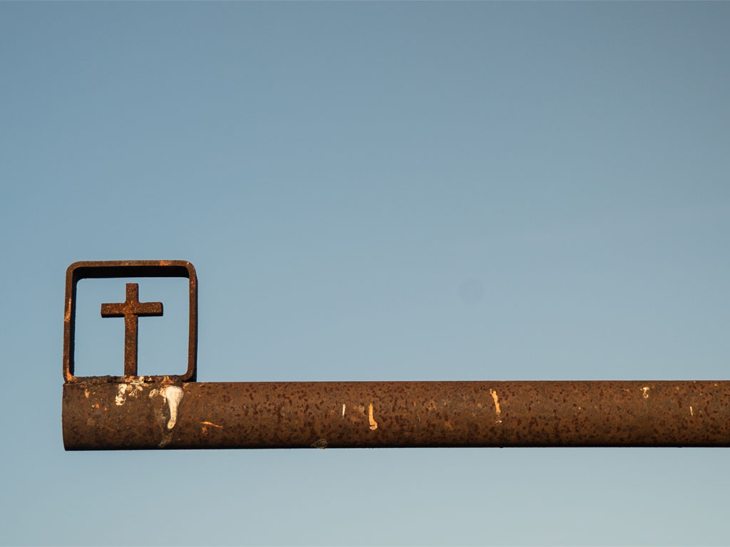 A long, rusted metal pole fills the screen entering from the right. On the far left side of the pole, a square with a cross is attached.
