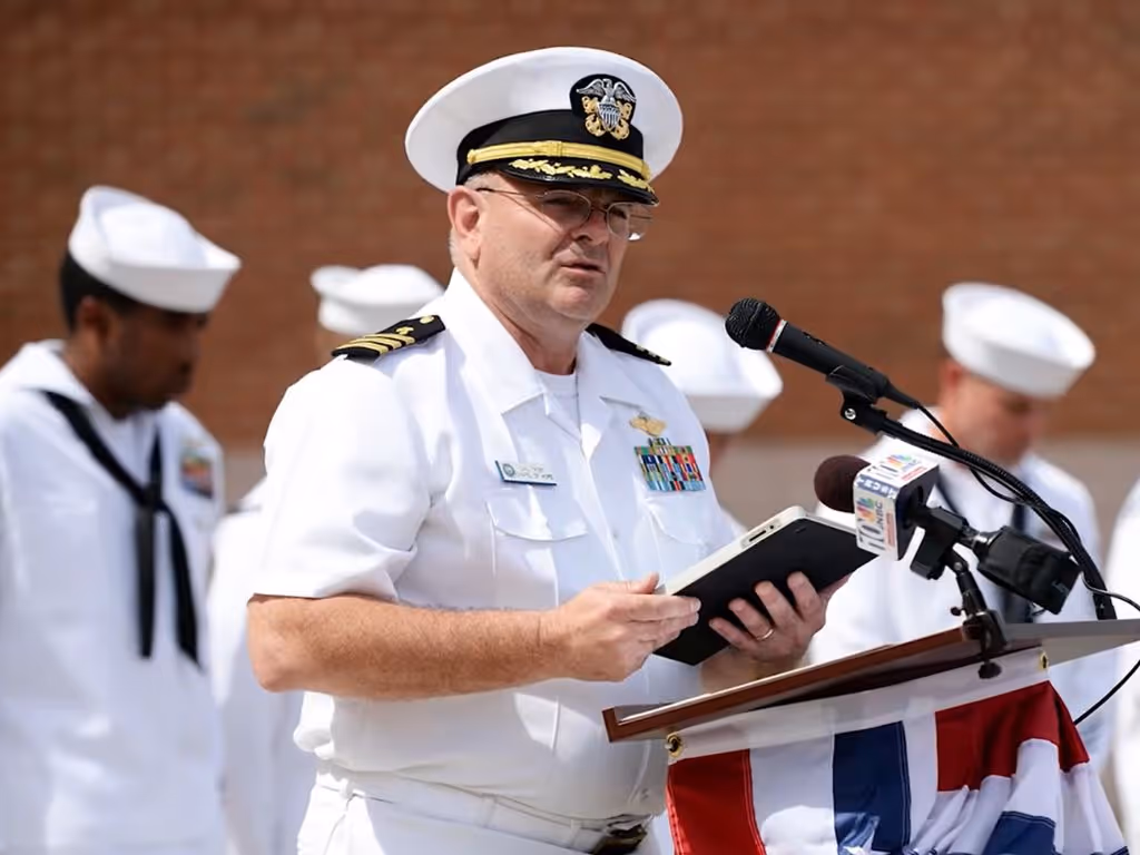 Person in military uniform speaking at a podium while holding a book, with others in uniform behind them