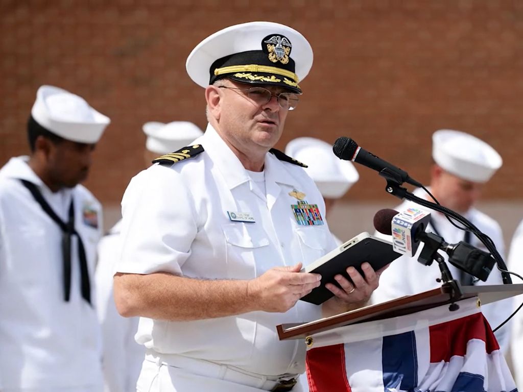 Person in military uniform speaking at a podium while holding a book, with others in uniform behind them