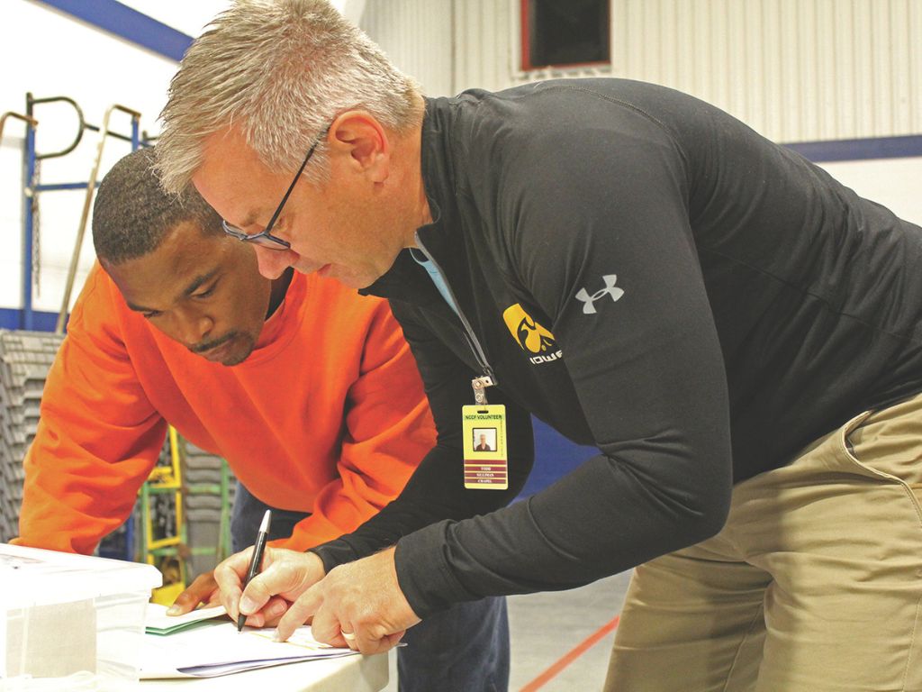 Man wearing a black sweatshirt leans over a table as he writes on a paper as a man in an orange sweatshirt watches closely.