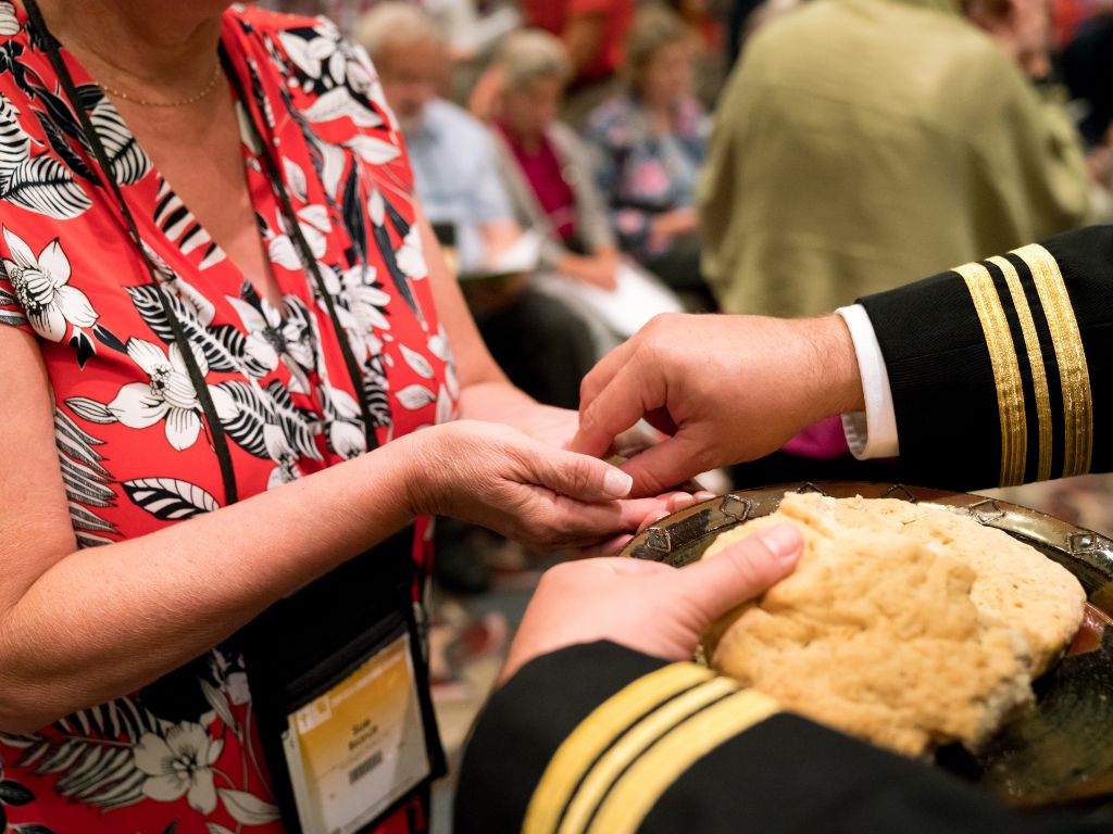 Hand of a person wearing a formal black military uniform places a piece of communion bread into the hands of a female wearing a red floral blouse. 