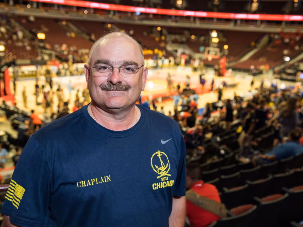 Man in a t-shirt that has “chaplain” on the righ chest smiles at the camera as he stands in an arena with an event happening in the background.