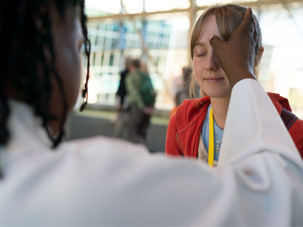 Person in a white robe makes the mark of the cross on the forehead of a female wearing a red sweater.