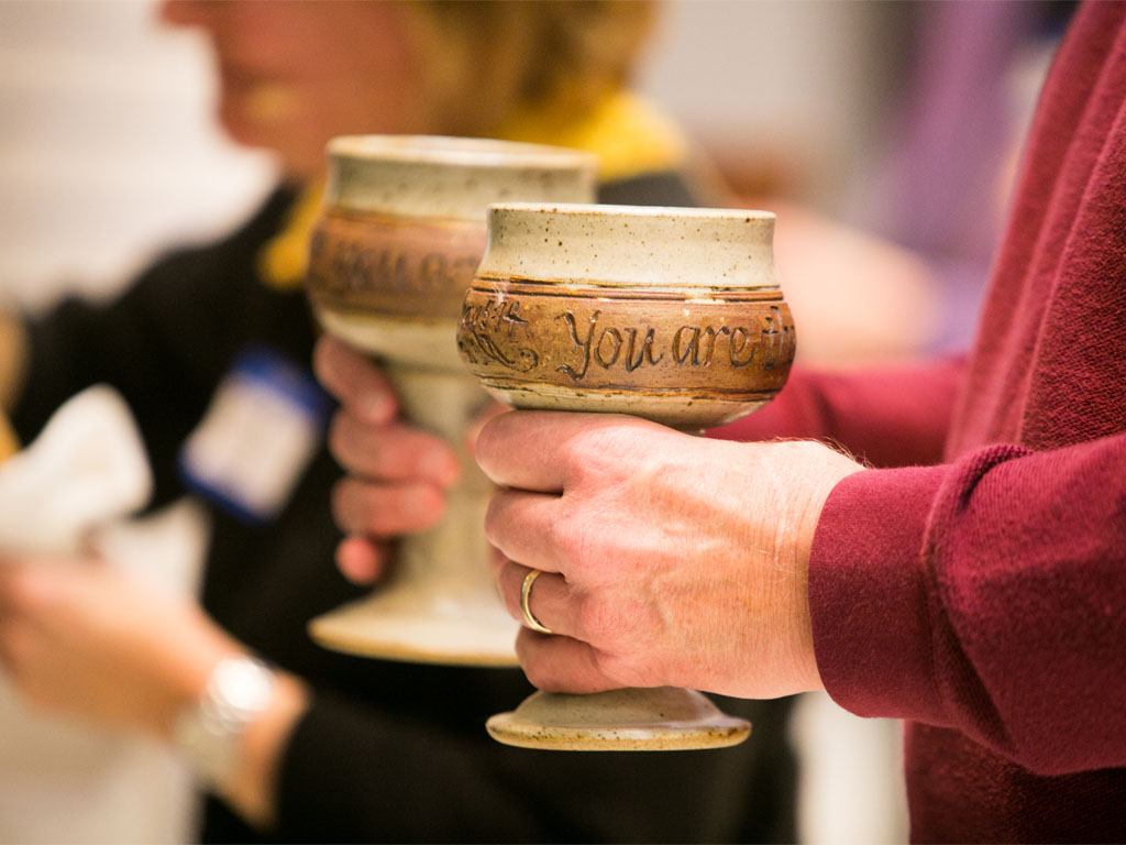A person with a red sweater whose face is not visible holds two clay chalices, one in either hand. The one closest to the camera has the inscription “You Are.”   