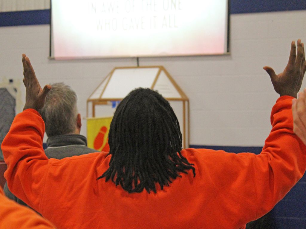 Person in orange jumpsuit with braided hair holds their hands up in praise as they look away from the camera.