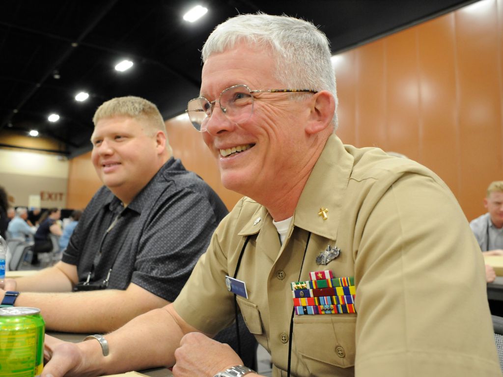 Man with glasses and short white hair wearing a tan military uniform with pins on the left pocket sits at a table smiling at something off camera.