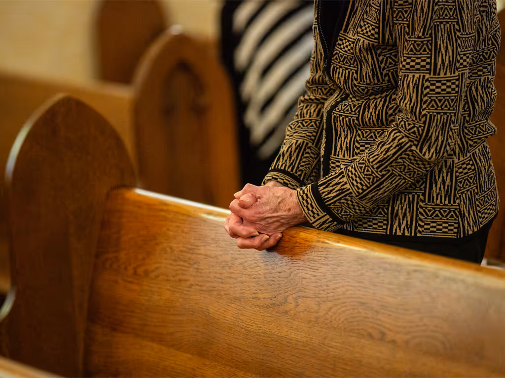 A person, whose face is off-camera, wearing a gold and black zip-up sweater has their hands folded in prayer, leaning on an empty church pew in front of them.