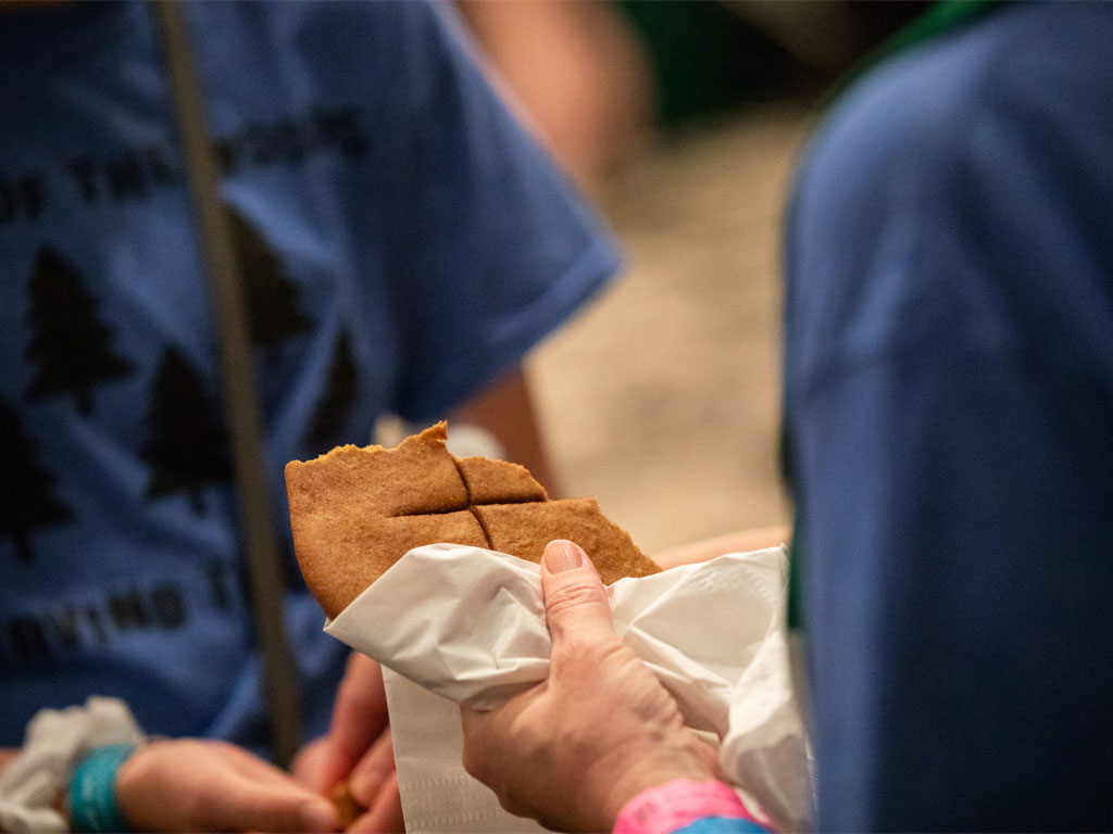 Figure in blue shirt holds a loaf of unleavened bread with a cross on it as another figure in a blue t-shirt holds a piece of bread in folded hands. 
