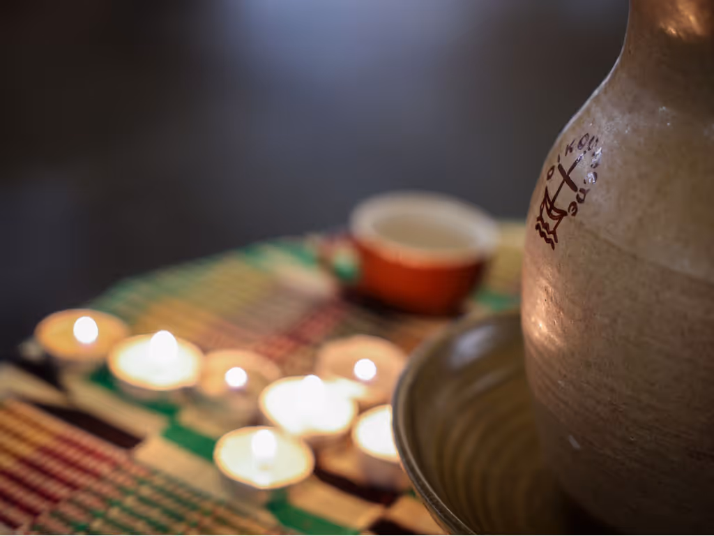 A pottery chalice with a cross and the word “oikoumene” on a table where out-of-focus votive candles are lit in a cross pattern. 