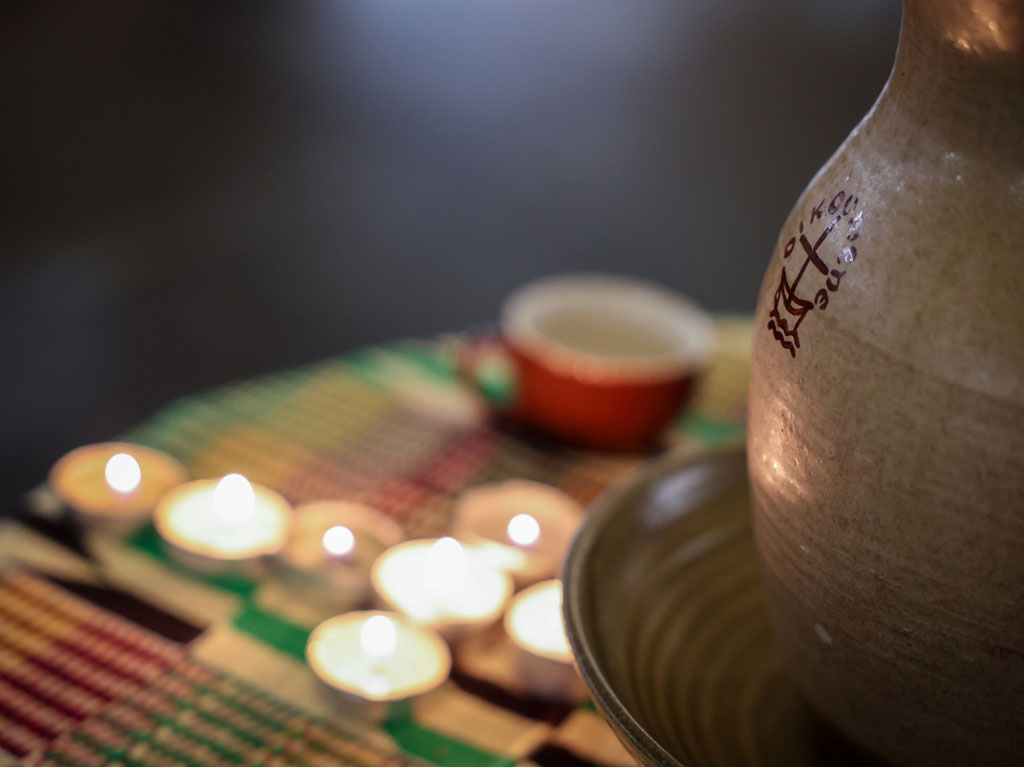 A pottery chalice with a cross and the word “oikoumene” on a table where out-of-focus votive candles are lit in a cross pattern. 