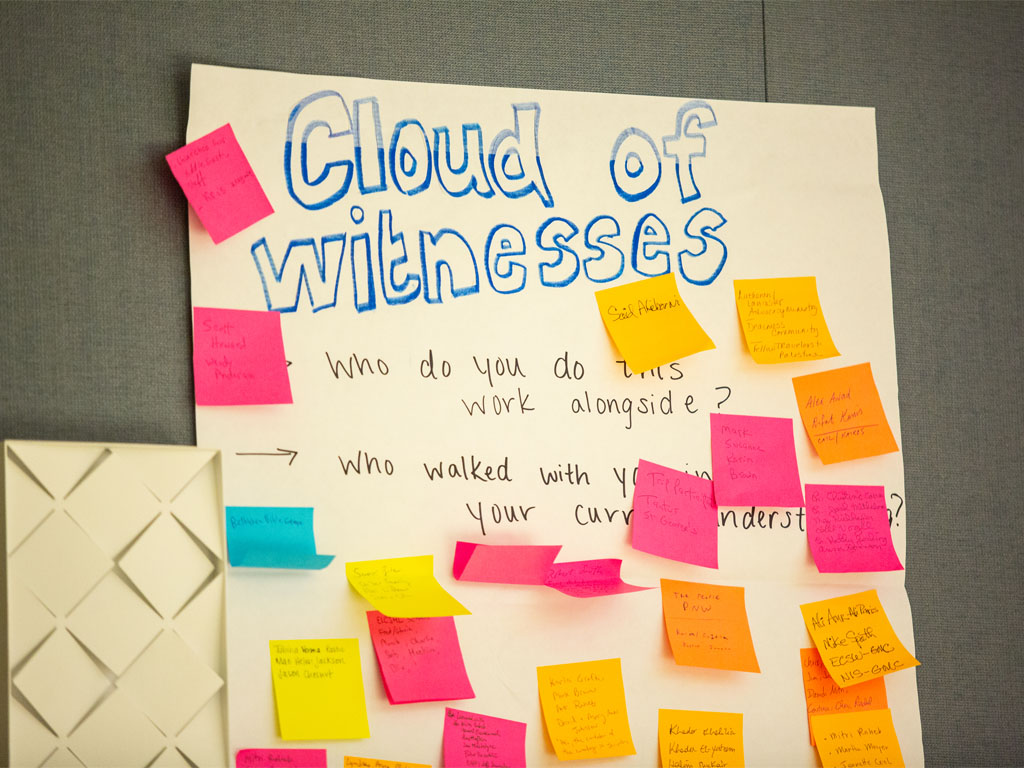 A piece of posterboard with the words “Cloud of Witnesses” and the questions “Who do you do this work alongside?” and “Who walked with you?” The board is covered with yellow, pink, orange and blue sticky notes.  