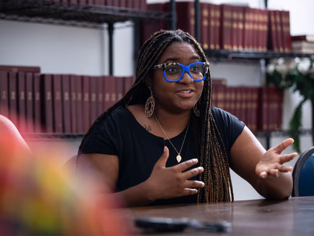 A woman with braids and blue glasses, wearing a black shirt, gestures and talks to people off-camera while seated in front of rows of purple books.