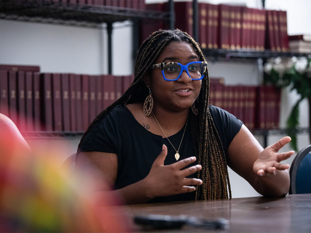 A woman with braids and blue glasses, wearing a black shirt, gestures and talks to people off-camera while seated in front of rows of purple books.