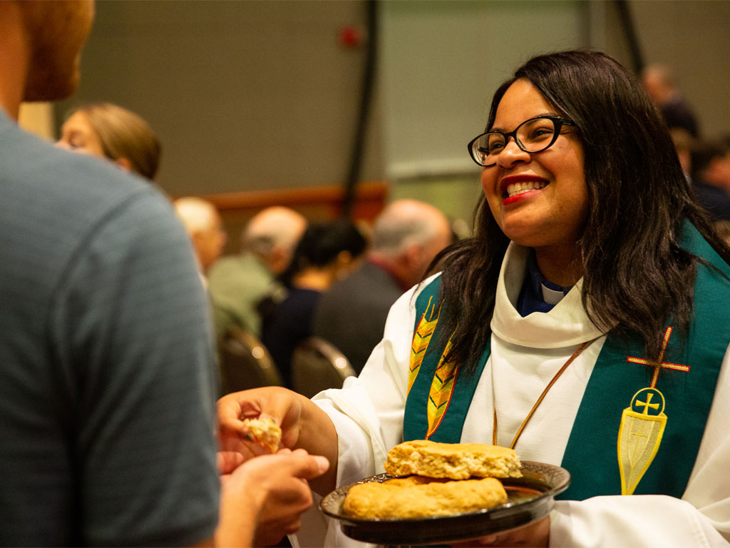 Female pastor wearing a white robe and green vestments smiles as she hands  a piece of communion bread to a man looking away from the camera wearing a gray short-sleeve shirt. 