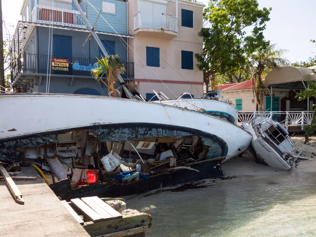 A long white boat sits on its side with a gaping hole in the hull showing large amounts of debris and trash after a storm.