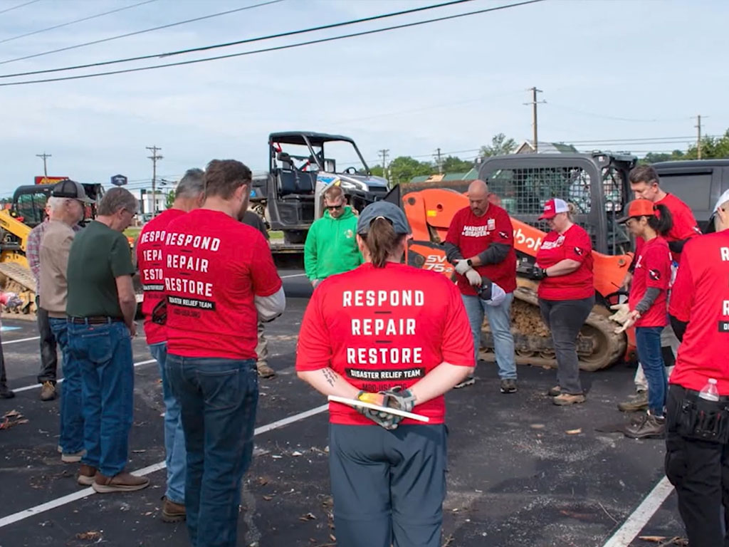 People standing in a circle in a parking lot bow their heads in prayer. They are standing in front of two skid loaders with many of the paricipants wearing red shirts that read “respond, repair, restore.”