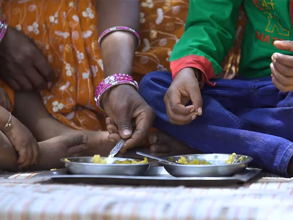 A mother's hand reaches in with a spoon into a metal bowl as she sits cross-legged with her children on a blanket. 