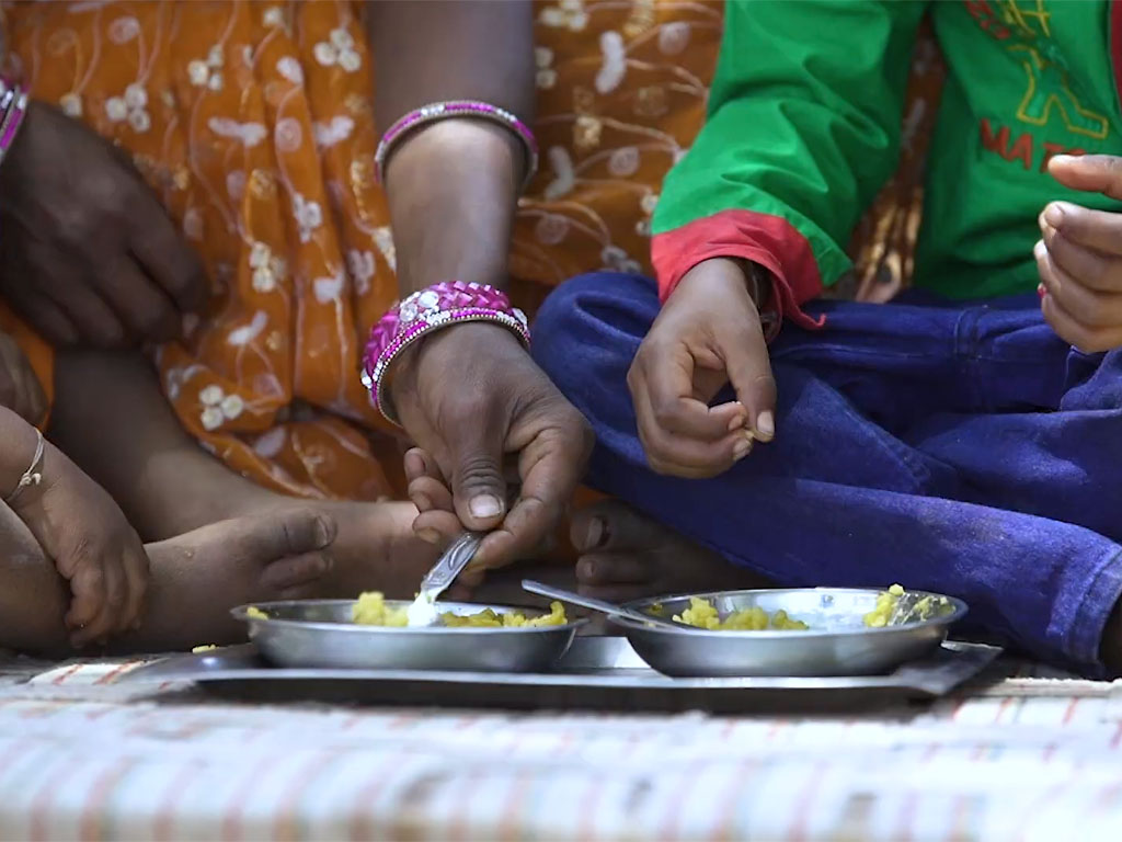 A mother's hand reaches in with a spoon into a metal bowl as she sits cross-legged with her children on a blanket. 