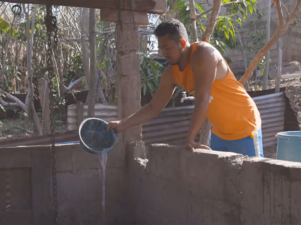 A man in an orange tank top pours water from a blue dish into a well. 