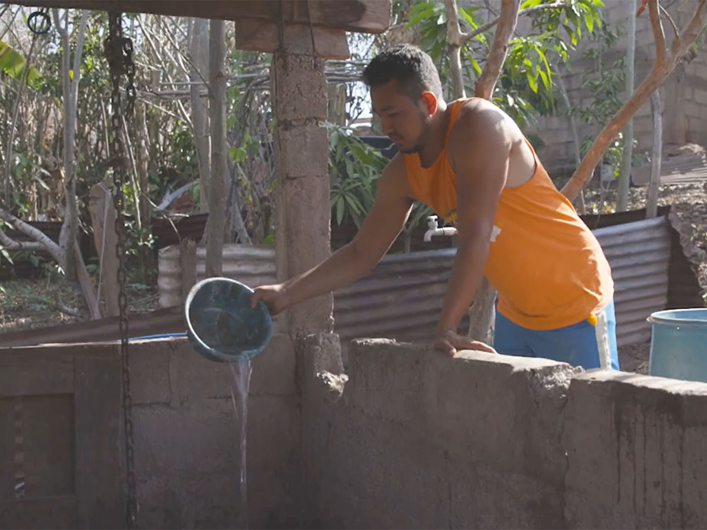 A man in an orange tank top pours water from a blue dish into a well. 