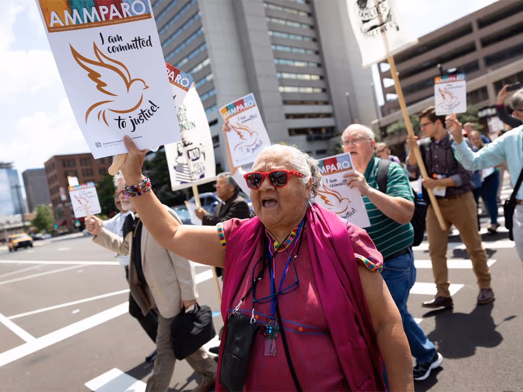A woman in purple, wearing red sunglasses, holds a sign that reads “AMMPARO I am committed to justice” as she leads a group of people crossing a city street.