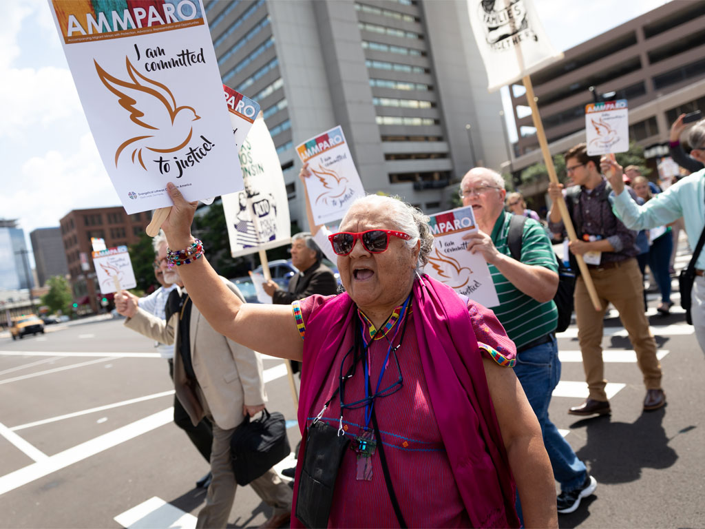 A woman in purple, wearing red sunglasses, holds a sign that reads “AMMPARO I am committed to justice” as she leads a group of people crossing a city street.