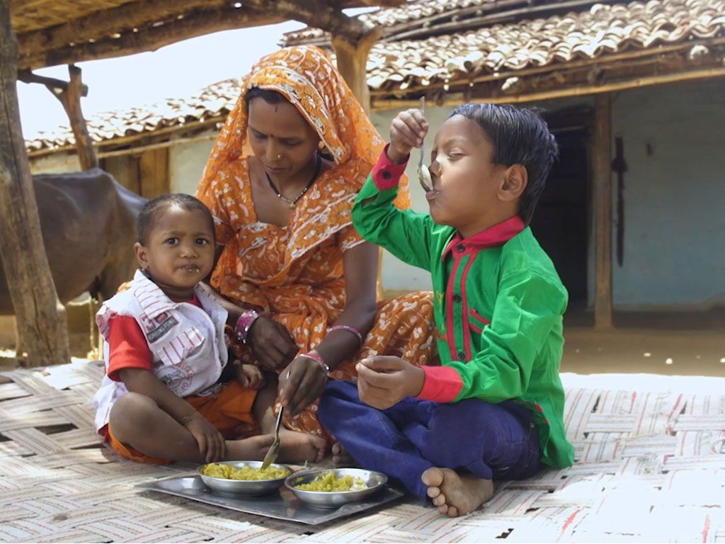 A mother wearing an orange dress and matching headscarf sits with two children as they eat green chutney out of metal bowls. 